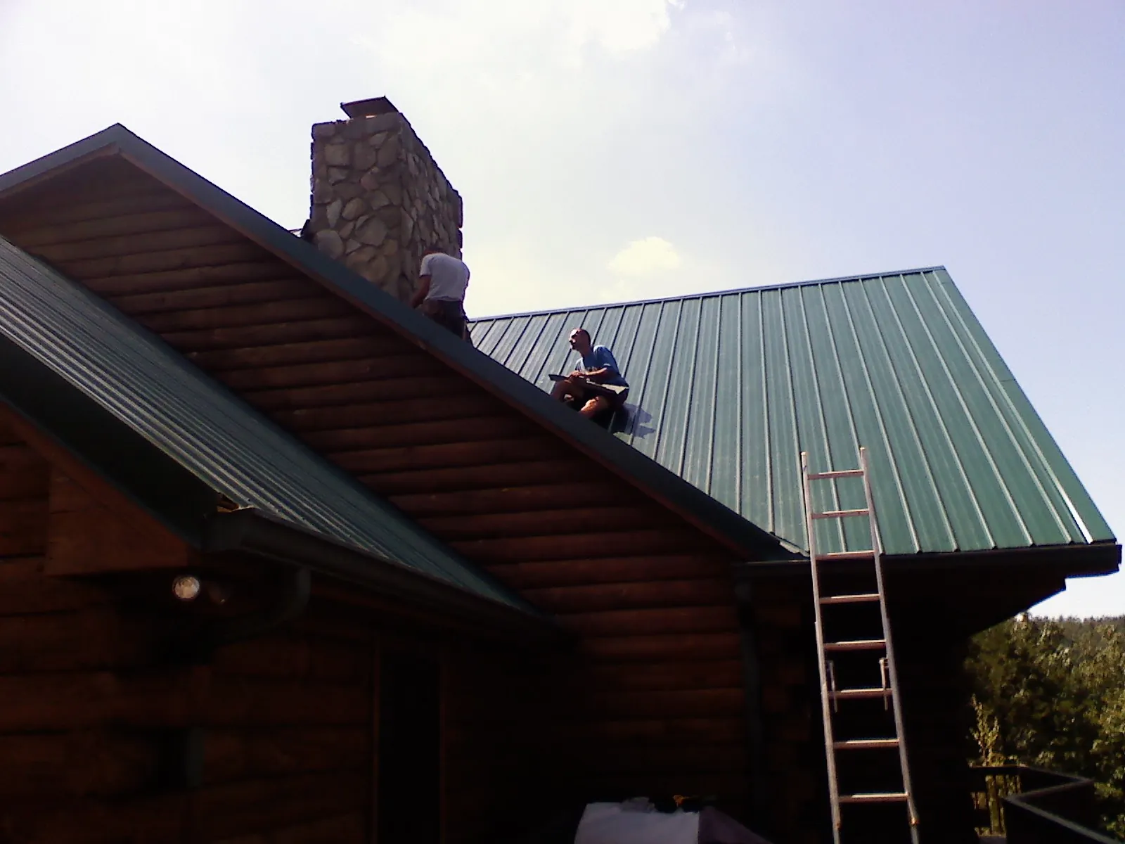 Roofers installing green metal panels beside a stone chimney on a log home in Kankakee